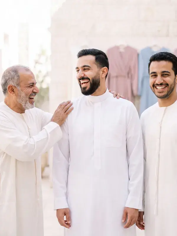 A groom in a white thobe laughing, his father's hand on his shoulder